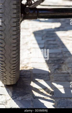 Detailed close-up of an old rubber tire with classic tread pattern on a textured cobblestone road. Strong sunlight casts sharp shadows from the vehicl Stock Photo