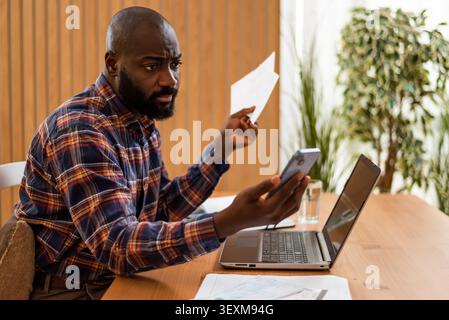 Holding an invoice in one hand and a smartphone in the other, a man carefully reviews his financial documents from home. Stock Photo