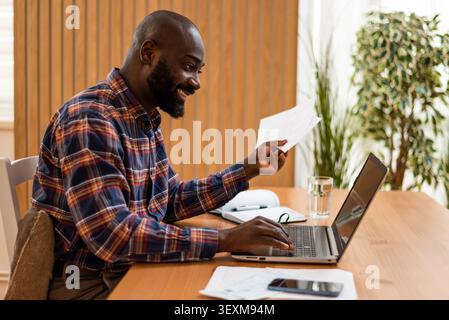 Black man compares paperwork with details on his laptop while working at home. Stock Photo