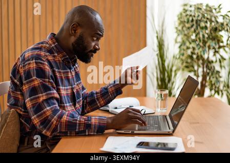 Black man compares paperwork with details on his laptop while working at home. Stock Photo