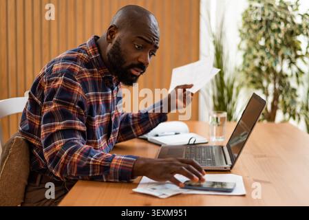 A concerned man sits at his desk reviewing bills while checking information on his smartphone. With a laptop open beside him, the scene illustrates th Stock Photo