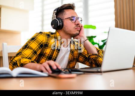 A stressed young man works on his laptop, resting his head in his hand, showing signs of frustration or mental fatigue. Stock Photo