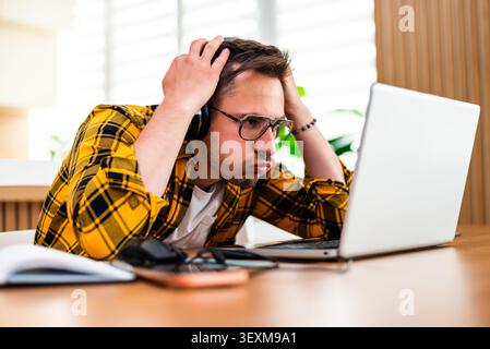 A stressed young man works on his laptop, resting his head in his hand, showing signs of frustration or mental fatigue. Stock Photo