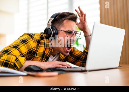 A man in headphones expresses visible frustration while working at his laptop in a home office. Stock Photo