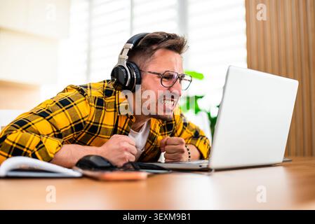A frustrated young man wearing headphones leans toward his laptop with clenched fists, reacting intensely to what he sees on the screen. Stock Photo