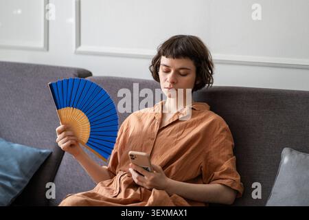 Tired woman using paper fan to cool down in room in summer, using mobile phone. Lack of ventilation Stock Photo