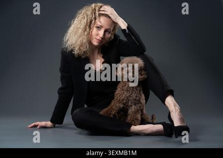 A young woman sits on the floor of a dark studio with a brown toy poodle leaning into her side. Their relaxed posture creates a stylish and modern Stock Photo