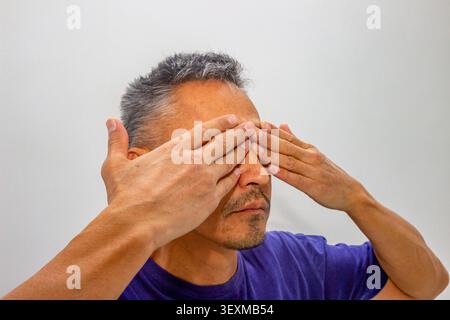 Mature Hispanic man covering his eyes with his hands. Concept of ignoring reality, denial and visual stress. Exhausted individual experiencing fatigue Stock Photo