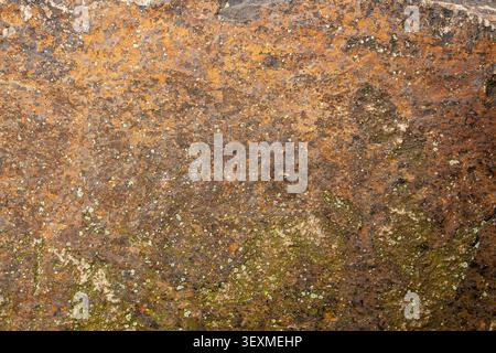 A rough, granular basalt stone surface displays warm orange and brown mineral staining with patches of green lichen and moss, creating a natural, weat Stock Photo