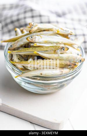 Fresh small fish stacked in a clear glass bowl on a white kitchen table, ideal for seafood recipes, cooking prep, market produce and healthy protein m Stock Photo
