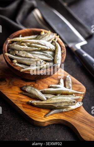 Fresh small fish arranged on a wooden cutting board with a rustic bowl blurred in the background, suggesting seafood market freshness, cooking prepara Stock Photo
