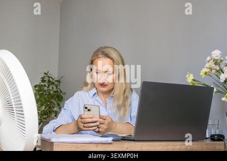 Woman checking a smartphone while working beside a running pedestal fan and laptop in a home office. Concept of connected living, smart home technolog Stock Photo