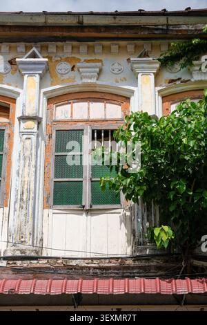 Architectural detail of a historic colonial-era shophouse featuring an arched wooden window, decorative plasterwork, weathered paint and tropical gree Stock Photo