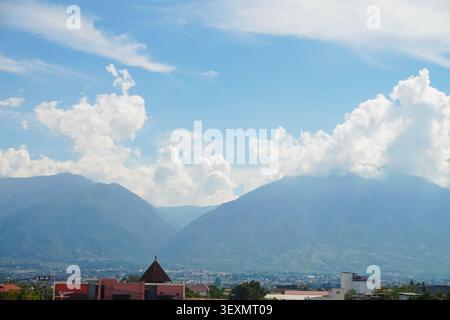 Vibrant clouds drift above distant mountains. Red-roofed buildings nestle at valley's edge. Majestic peaks frame the tranquil landscape. Sky's soft blue hints at peaceful calm Stock Photo
