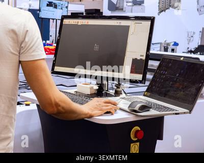 An operator uses specialized CAD/CAM software on a dual-screen computer setup to control an industrial CNC cutting machine in a modern manufacturing f Stock Photo