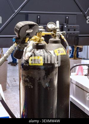 Two high-pressure black metal gas cylinders equipped with brass valves, regulators, and pressure gauges, set up in an industrial or exhibition environ Stock Photo