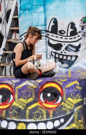 Young woman knitting amid vibrant street art murals reflecting Amsterdam's creative urban culture, Netherlands Stock Photo