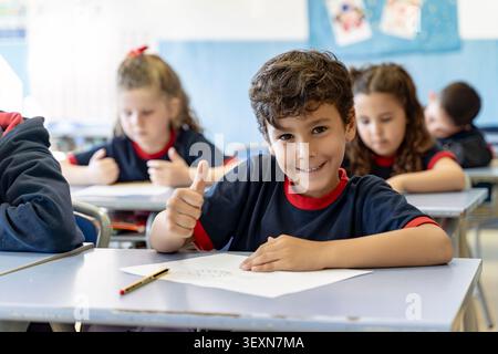 Smiling elementary school boy student showing thumbs up gesture, sitting at desk in a bright classroom with classmates Stock Photo