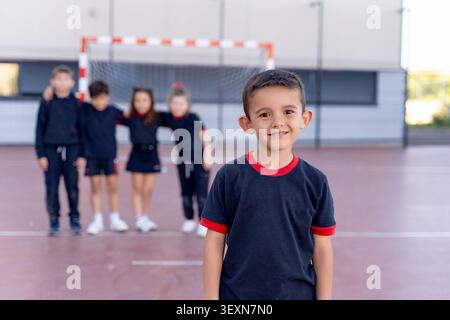 Schoolboy smiling at camera during physical education class. Classmates standing with a goal and court in background Stock Photo