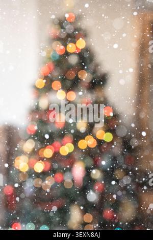 Vertical festive background featuring colorful Christmas tree bokeh lights in a warm home interior. A soft overlay of falling snowflakes adds a magica Stock Photo