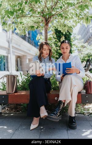 Two businesswomen collaborating outdoors on a bench with tablet and documents in city park Stock Photo
