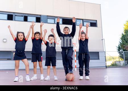 Diverse group of elementary students in school uniforms cheering with arms raised on a school playground, expressing joy and teamwork Stock Photo