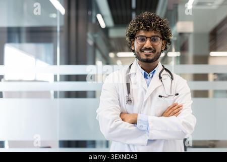 Young indian male doctor in white coat and stethoscope smiling at camera, standing with arms crossed in a modern hospital or clinic corridor, representing healthcare and medical professionalism Stock Photo