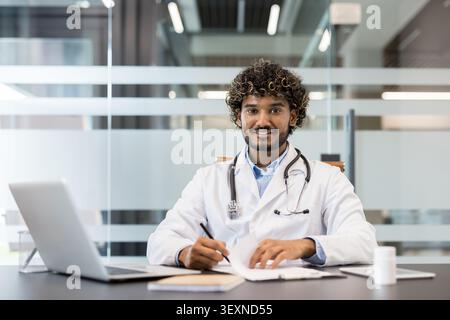 Young indian male doctor smiling, looking at camera while sitting at his desk, wearing a lab coat and stethoscope, writing notes in a bright, contemporary clinic office Stock Photo