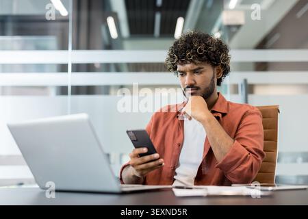 Young indian man with curly hair focusing intently on his smartphone while working at a desk with a laptop in a modern office, feeling thoughtful and concentrated Stock Photo