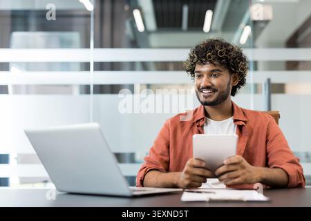 Indian man sitting at a desk, looking away from his laptop and holding a digital tablet, demonstrating a positive attitude while engaged in remote work Stock Photo