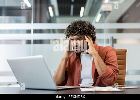 Young man experiencing burnout and pressure, feeling frustrated and stressed, holding his head while working on a laptop at a modern office desk, dealing with a headache Stock Photo