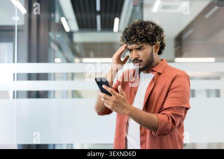 Indian man experiencing stress and worry, holding his hand to his head while looking at bad news on a mobile phone in a modern office environment Stock Photo