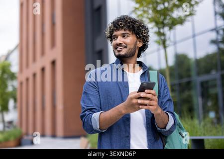Young man with curly hair and backpack walking on a college campus while looking away and smiling, using his mobile phone for communication and connection Stock Photo