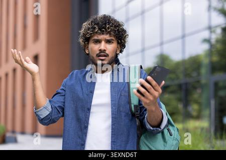 Young man standing outdoors, looking confused and stressed, shrugging with one hand up while holding a smartphone, experiencing navigation issues or a mobile problem Stock Photo