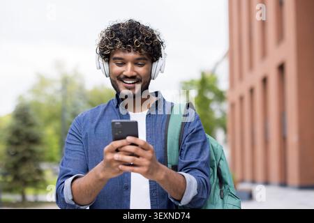 Happy indian student wearing headphones, actively using a smartphone, and carrying a backpack while walking on a university campus, representing modern higher education and connectivity Stock Photo