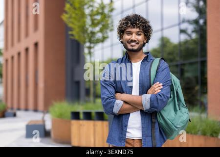 Young male university student with curly hair and a backpack standing outdoors on a modern campus, confidently smiling while crossing arms and looking at camera Stock Photo