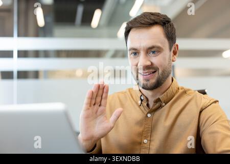 Man smiling and waving hand during a remote online video call using a laptop in a modern office creating a sense of connection and virtual communicati Stock Photo