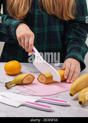 Child in a green plaid shirt cutting a banana with a safe kids plastic knife on a pink cutting board, learning to cook in the kitchen, close-up Stock Photo