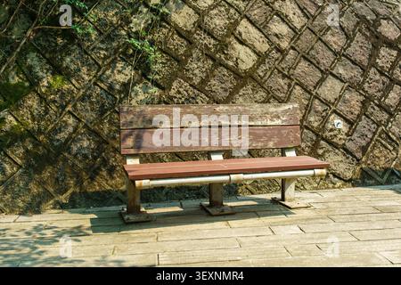 An old weathered wooden bench placed on a stone paved walkway against a structured stone retaining wall in outdoor sunlight. Stock Photo