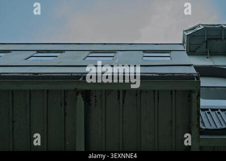 Low angle view of a pale green metallic industrial building with three rectangular roof windows against a soft cloudy sky. Stock Photo