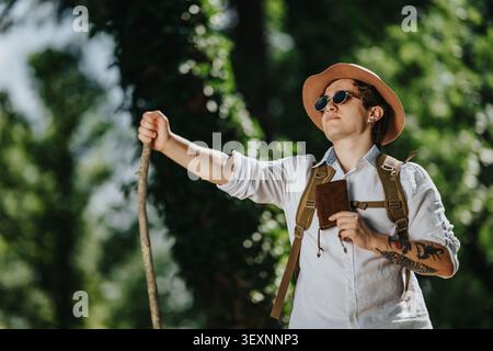 Young male hiker with backpack and notebook standing in forest holding walking stick and looking up Stock Photo