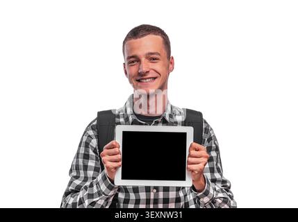 A smiling young male student with a backpack holds up a digital tablet with a blank black screen. Stock Photo