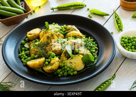 Healthy new potato salad with fresh peas, baby spinach, herbs to garnish, and  dressing  with mustard, vinegar, and oil in black plate on white wood b Stock Photo