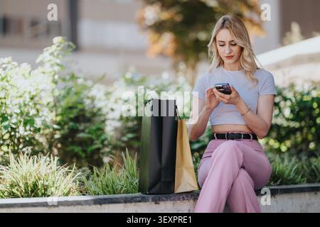 Young woman shopping outdoors sitting on a bench using smartphone with shopping bags Stock Photo