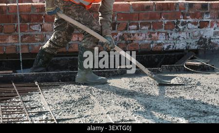 Worker Spreading Wet Concrete at Construction Site. Cropped construction worker in boots and gloves spreads wet concrete with a long handled shovel Stock Photo