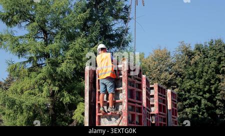 Worker Securing Red Concrete Formwork Panels. Construction worker in hard hat and orange vest stands on red concrete formwork panels, securing crane Stock Photo