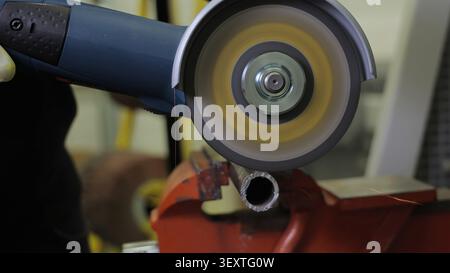 Angle Grinder Cutting Metal Pipe in Workshop. Close-up angle grinder with a spinning abrasive disc cuts a metal pipe held in a red vise, creating Stock Photo