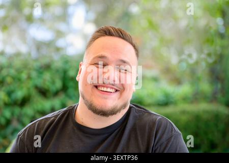 Happy young man smiling and laughing naturally while sitting outdoors in a relaxed environment. Authentic lifestyle portrait with positive emotion, fr Stock Photo