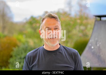 Friendly mature man smiling and winking outdoors in a relaxed natural environment. Authentic lifestyle portrait with casual clothing, positive emotion Stock Photo