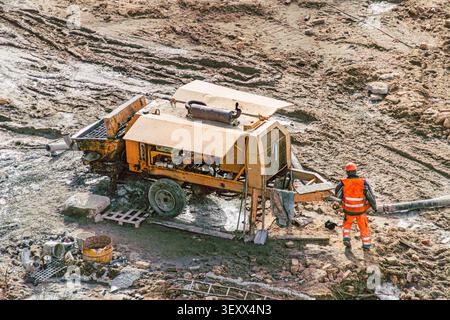 Construction site featuring heavy machinery, likely a concrete mixer or similar equipment, situated on muddy terrain. A worker in high-visibility vest Stock Photo